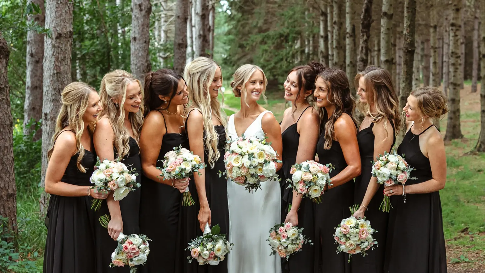 A bride and her bridesmaids in black dresses holding hand-tied bouquets of cream and blush blooms.